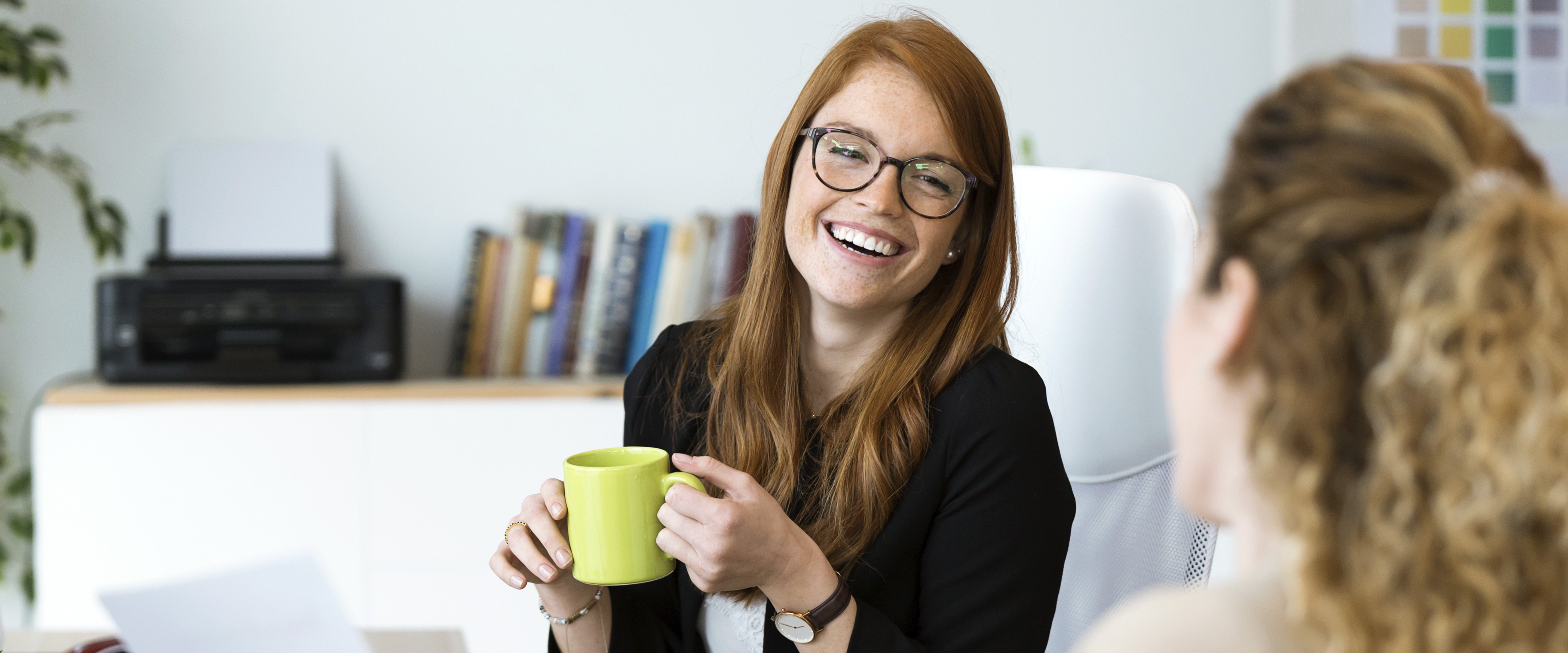 smiling woman with coffee cup talking to coworker at desk